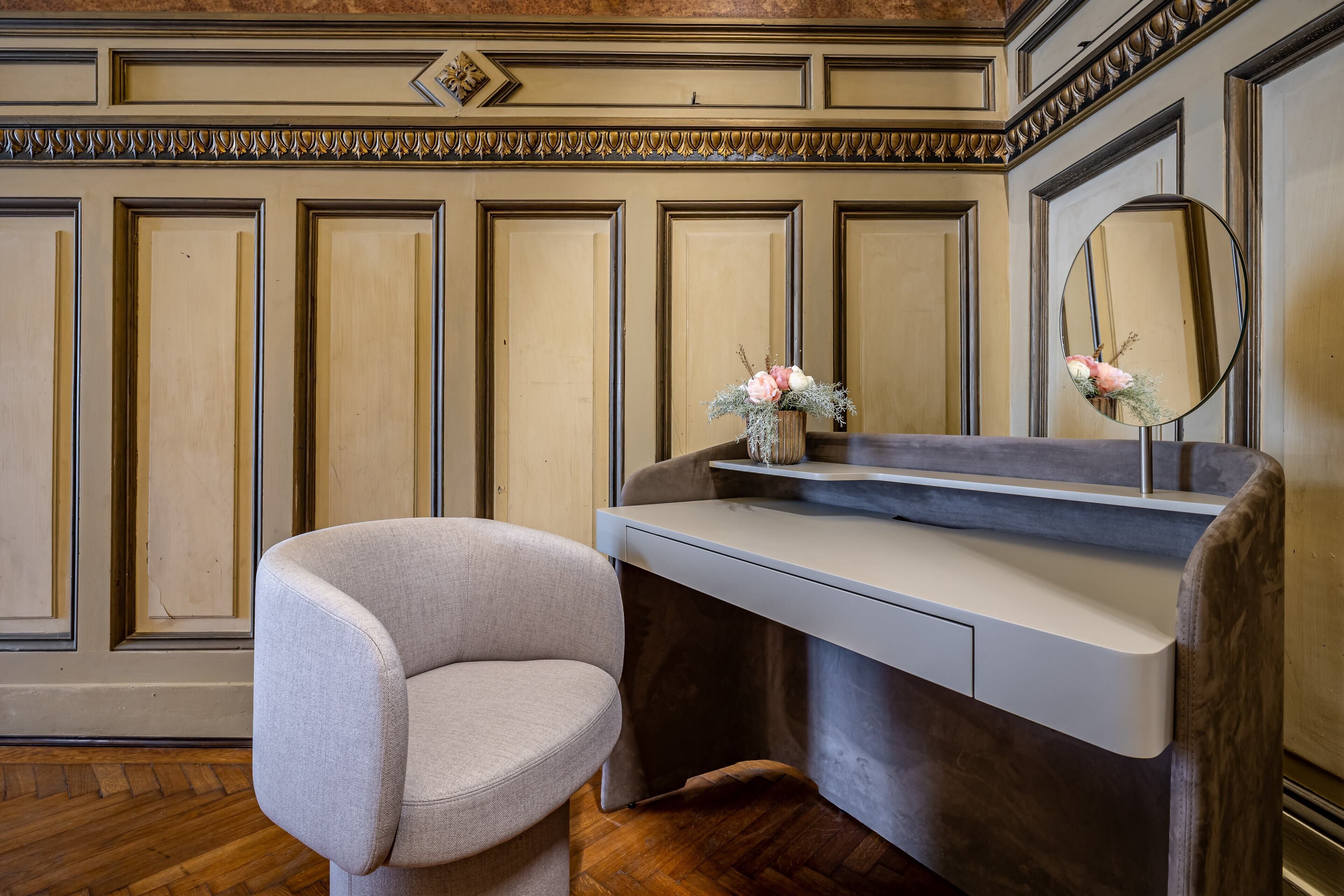 A vanity area with a chair, a mirror, and a vase with flowers.