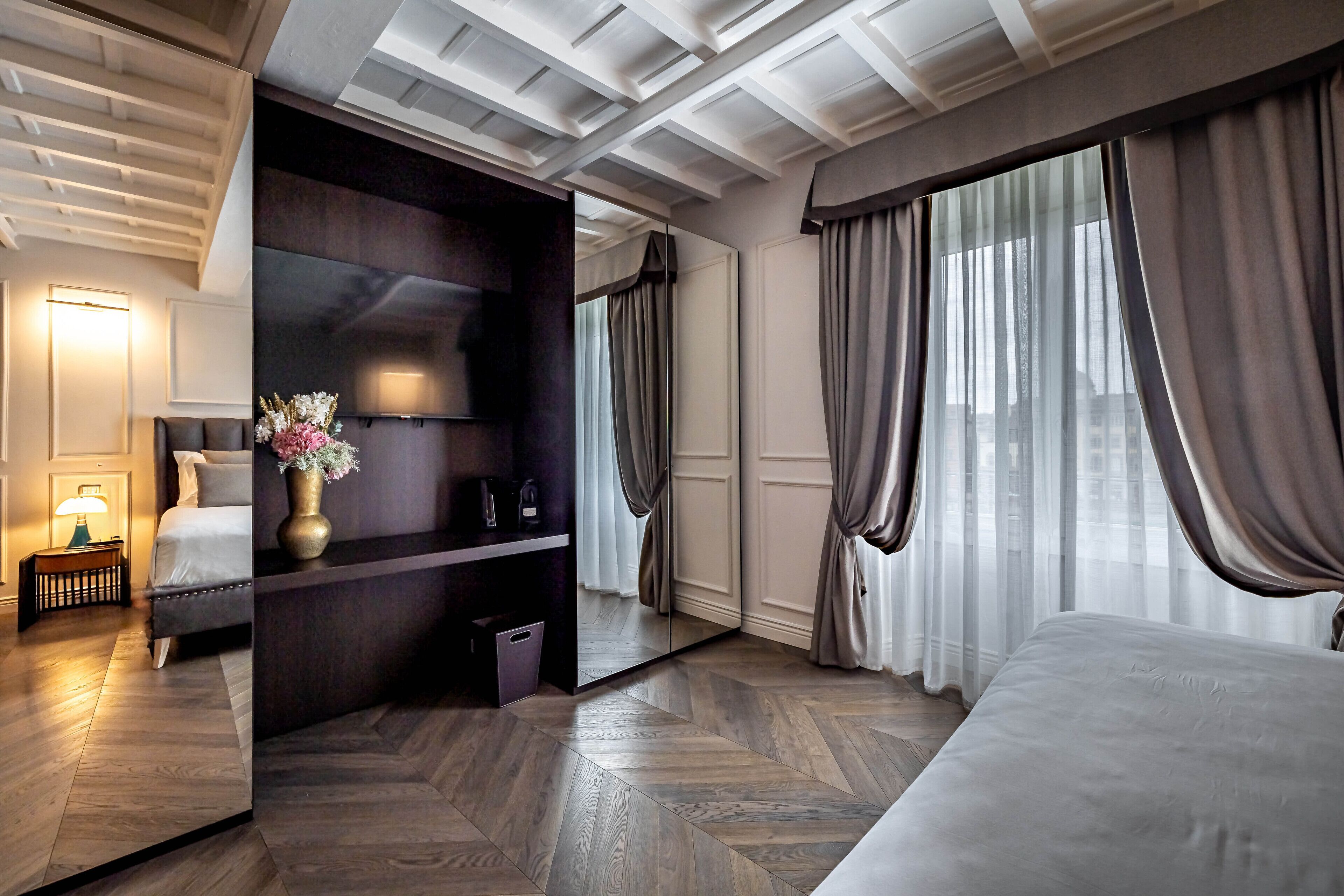 Una habitación de hotel moderna con una cama grande, un área de descanso con sofá y sillones, una mesita con lámpara, un espejo y un ventanal con cortinas.