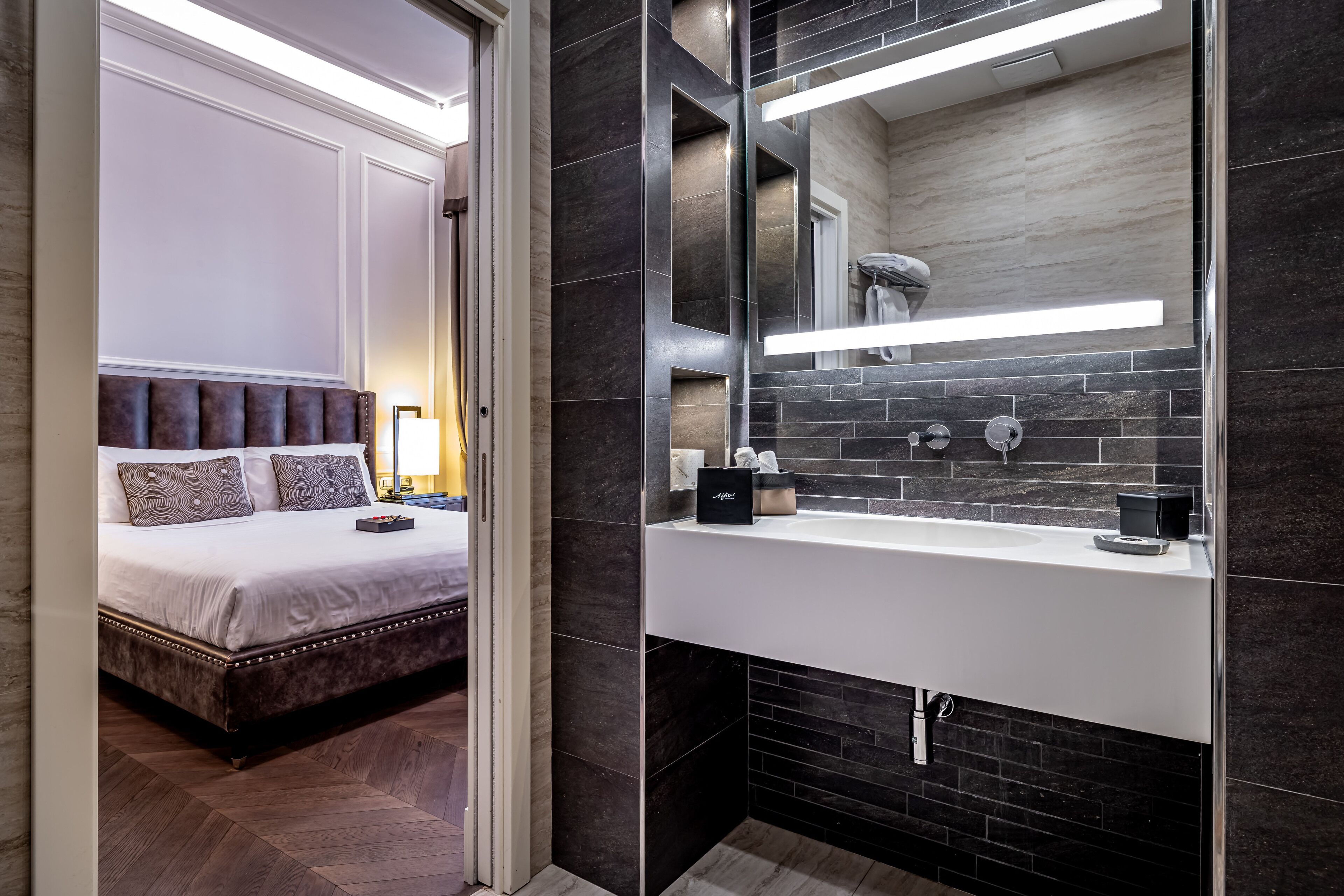 Ein modernes Hotelzimmer mit einem großen Bett, einem Badezimmer mit dunklen Fliesen und einem großen Spiegel.