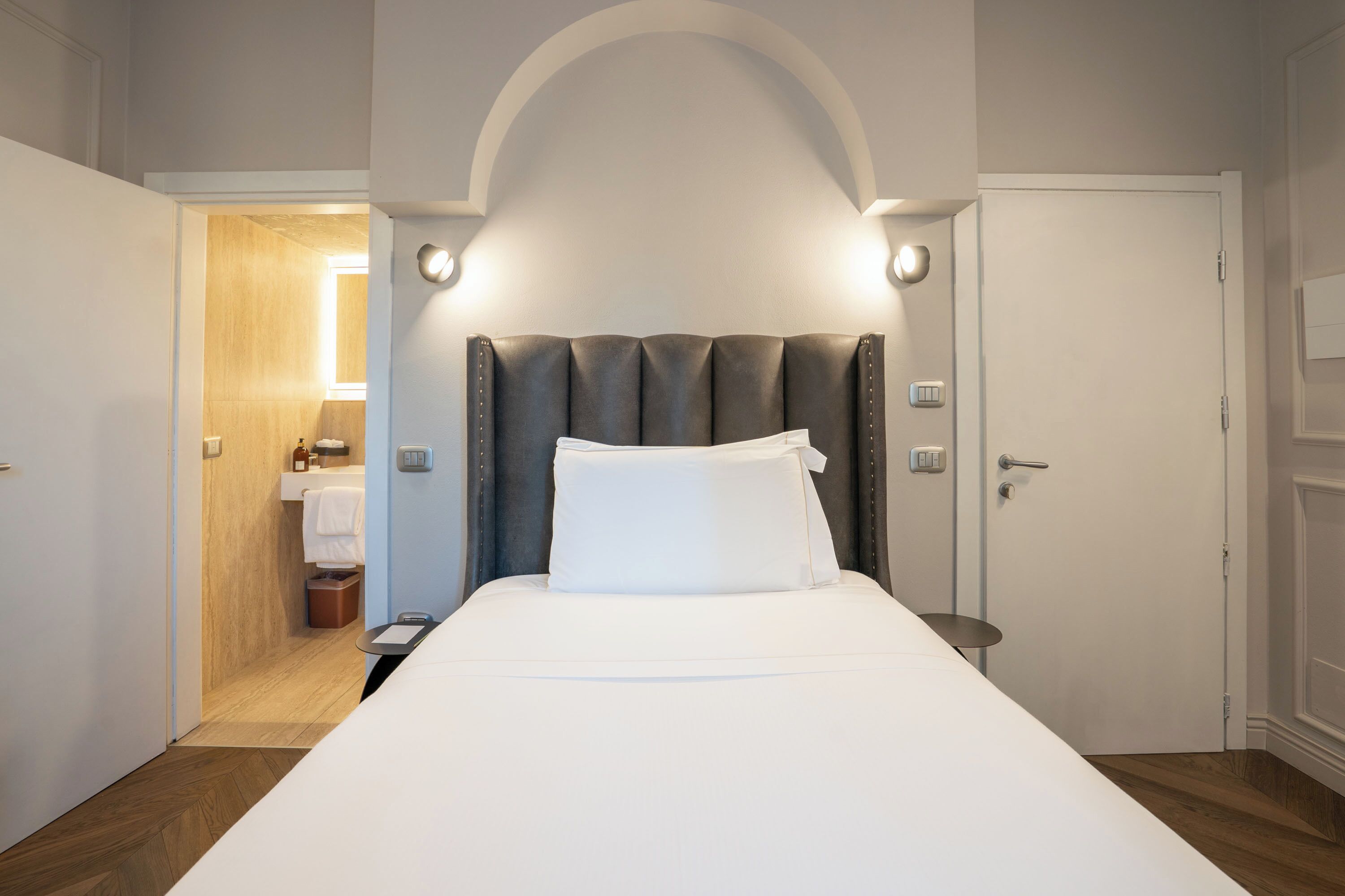 A hotel room with a bed, a bedside table, a nightstand, a door, and a bathroom visible through an open door.