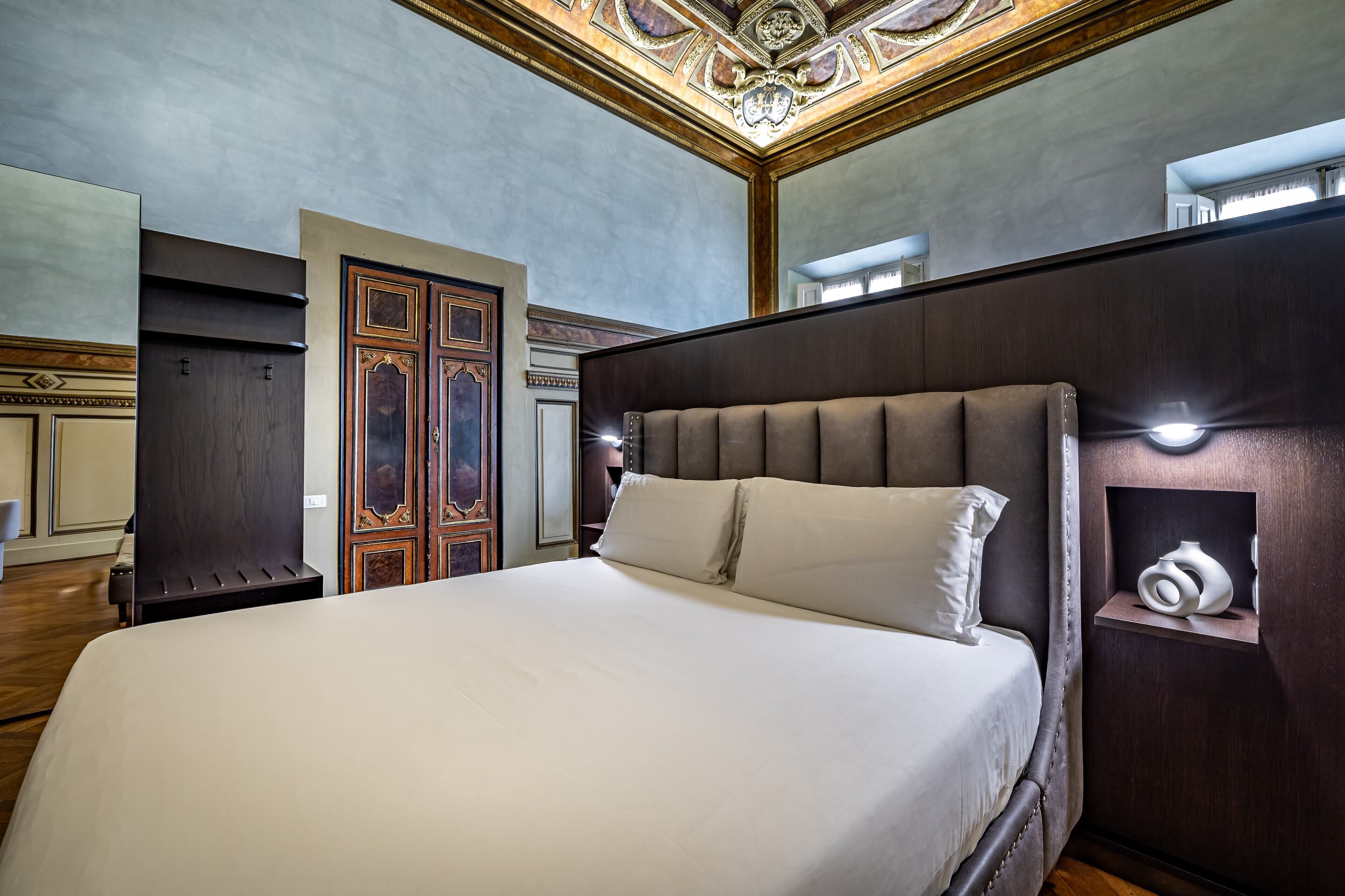 A hotel room with a large bed, a wooden wardrobe, a mirror, and a small shelf with a vase.