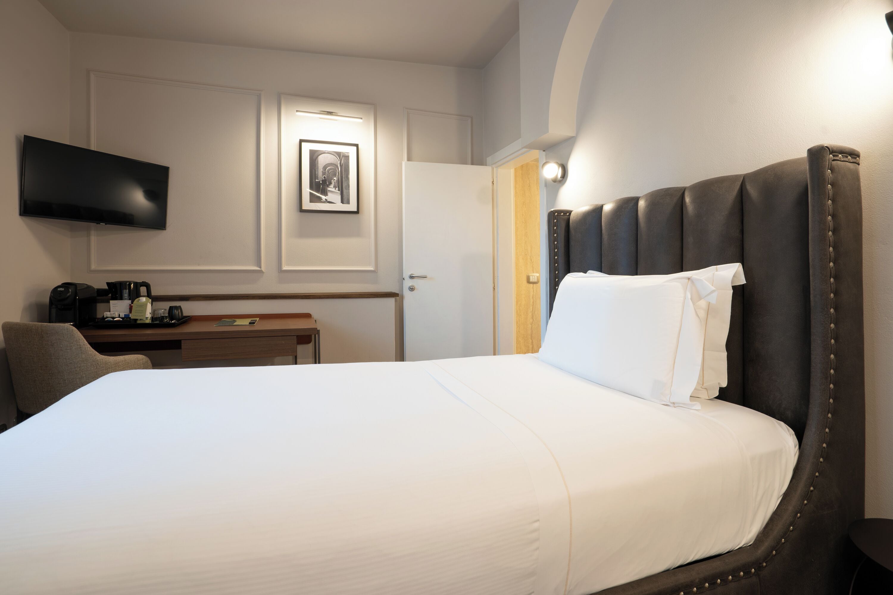 Hotellihuone, jossa on suuri sänky, seinälle asennettu taulutelevisio, työpöytä kahvinkeittimellä sekä ovi, joka johtaa toiseen huoneeseen.