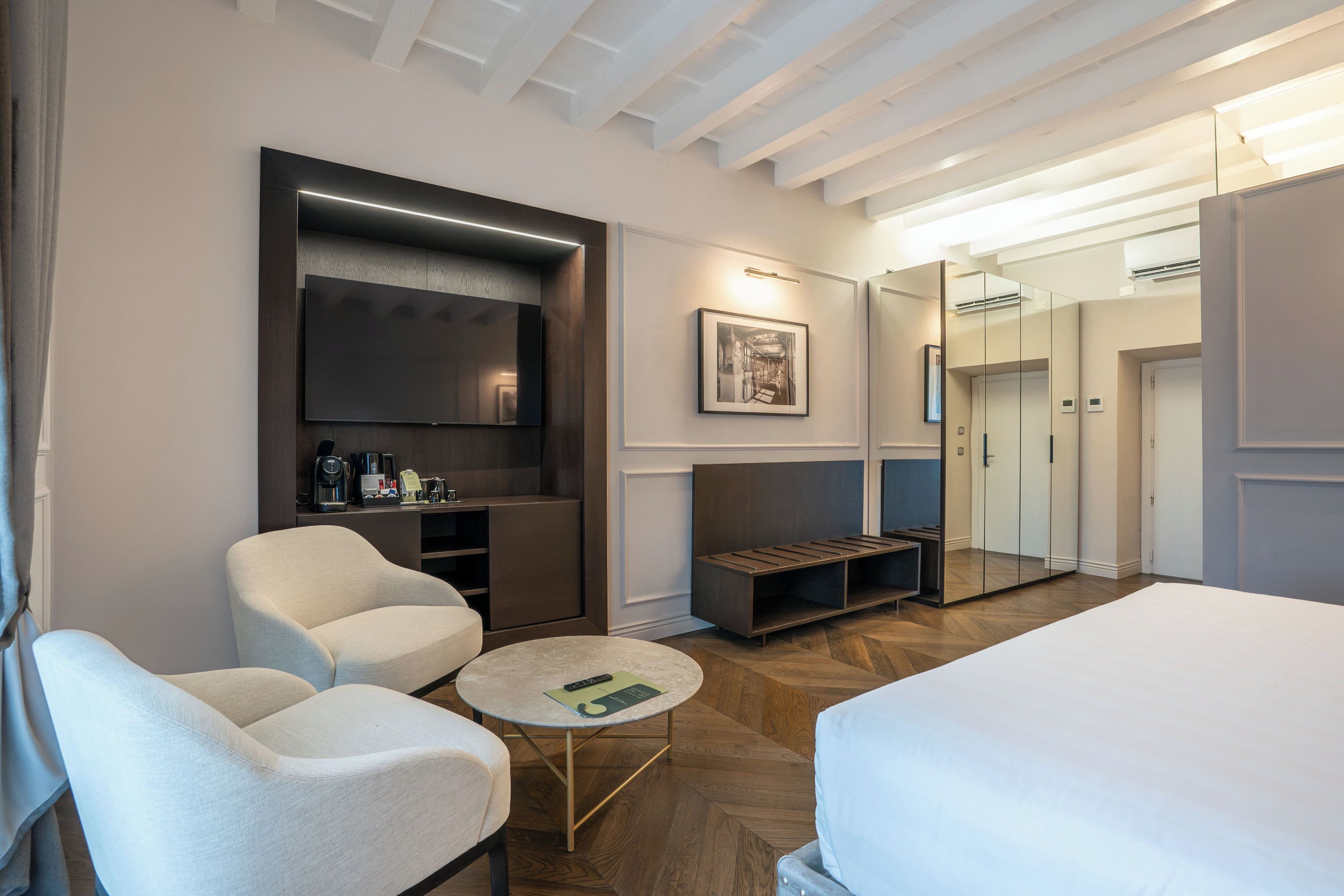Une chambre d’hôtel moderne avec un grand lit, deux fauteuils, une petite table ronde, un téléviseur à écran plat, une machine à café et un miroir.
