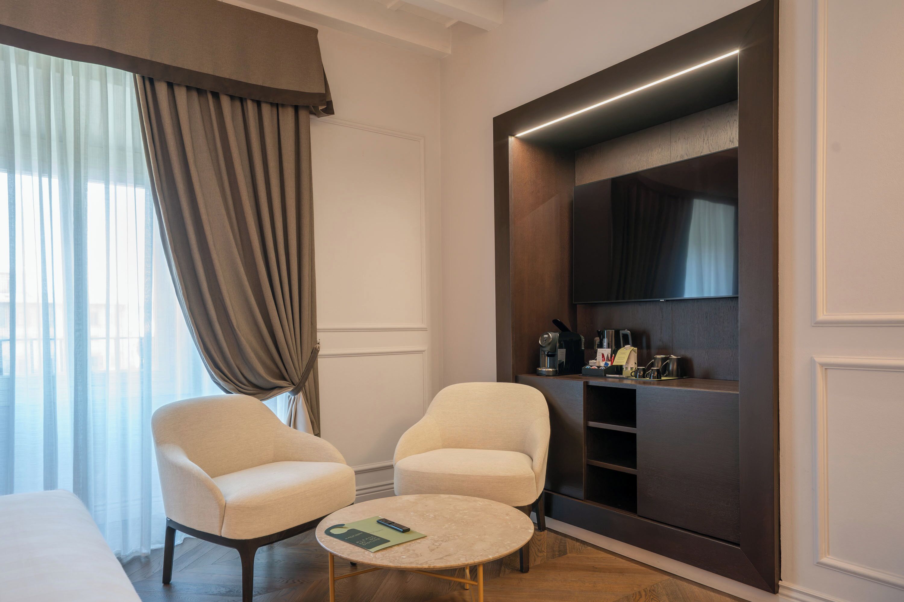 A hotel room with two armchairs, a small round table, a flat-screen TV, and a built-in shelf with kitchen appliances.