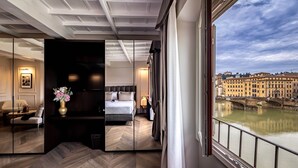 A modern hotel room with a large mirror, a bed, a seating area, and a view of a river and historic buildings. - Il Rivalta - Place of Charme (Florence)