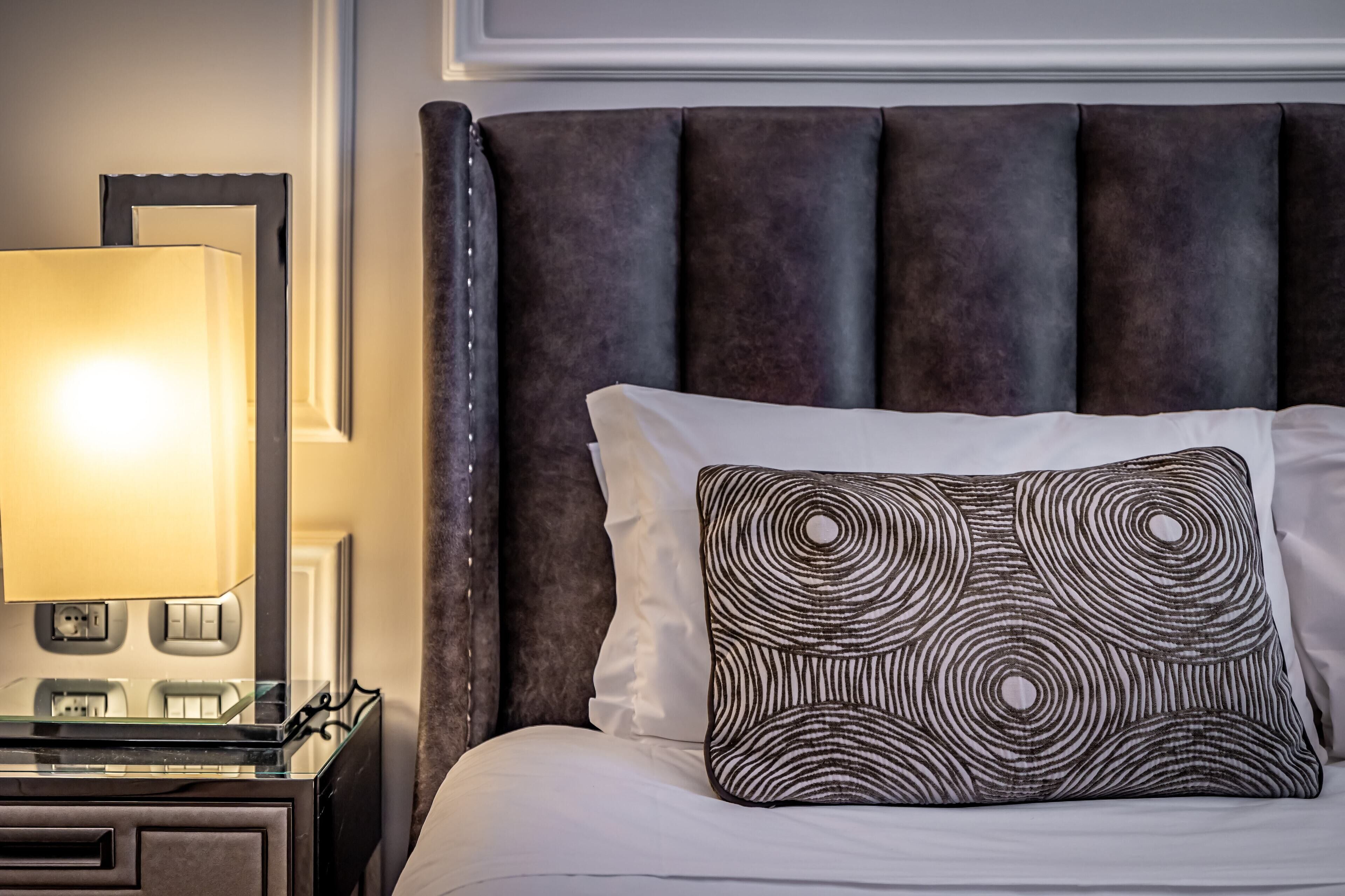 A neatly made bed with a patterned pillow, a bedside lamp, and a dark upholstered headboard.