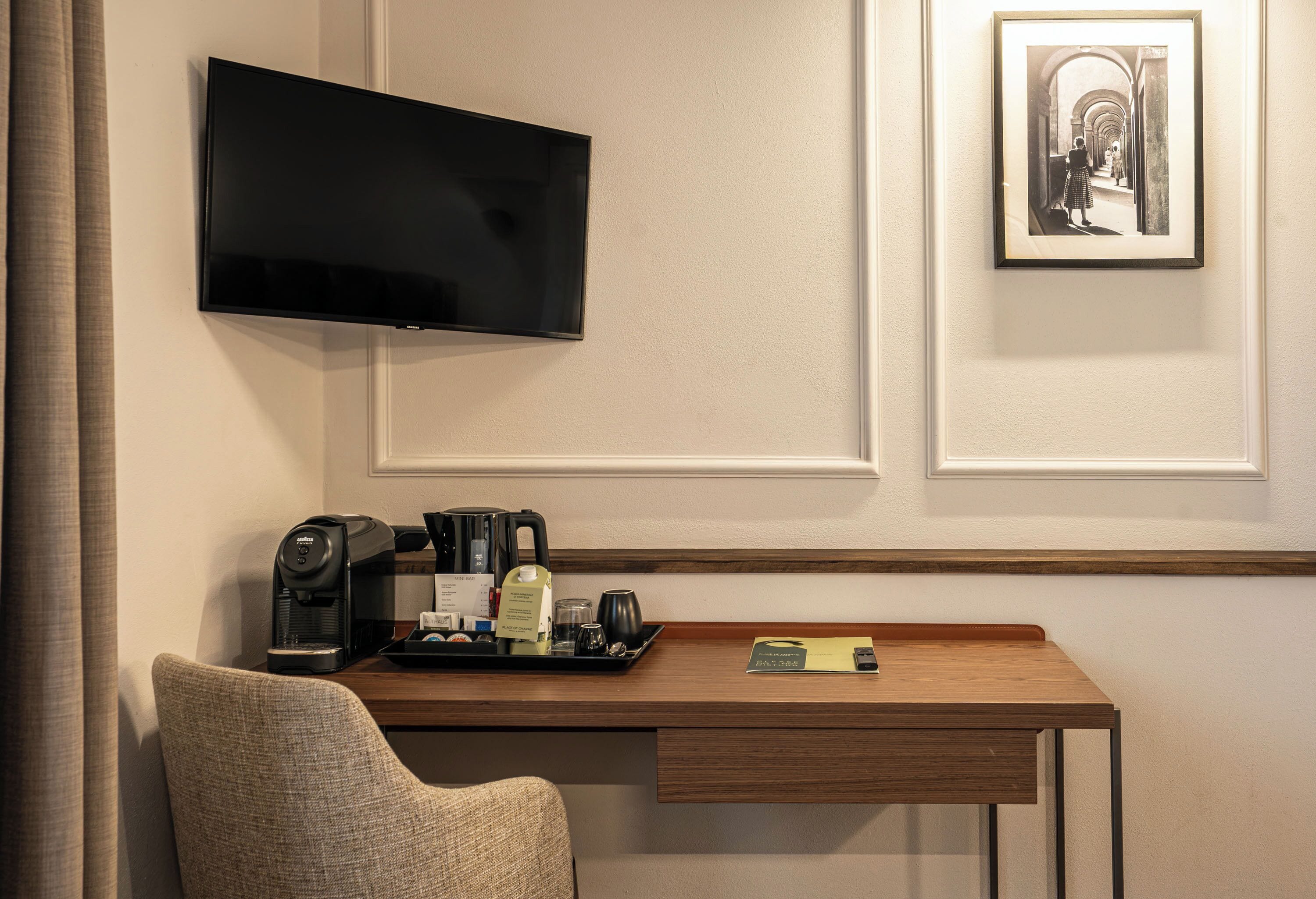 A hotel room with a flat-screen TV mounted on the wall, a coffee machine, a tray with a bottle and glasses, and a framed picture.