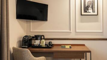 A hotel room with a flat-screen TV mounted on the wall, a coffee machine, a tray with a bottle and glasses, and a framed picture.