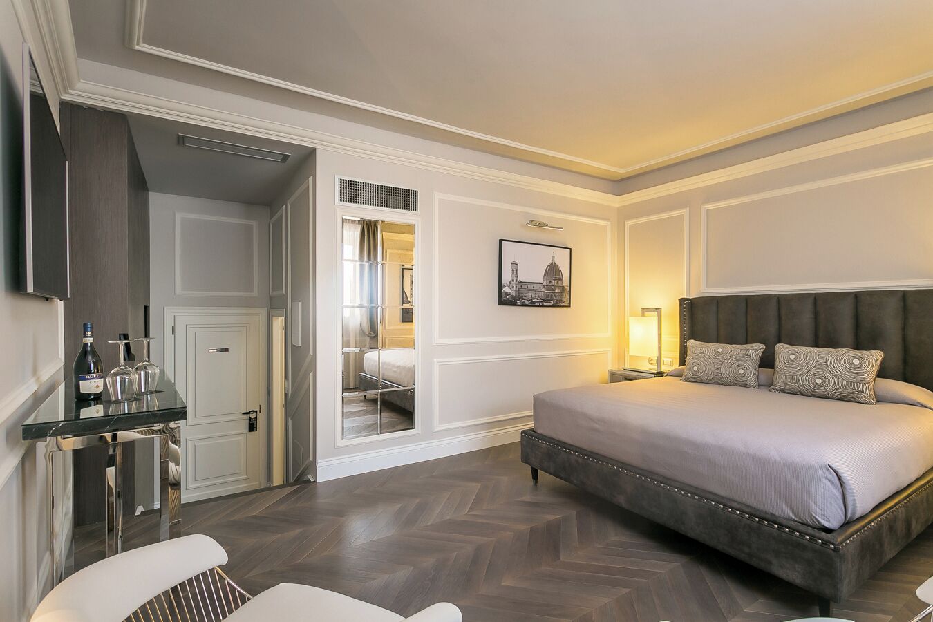 A hotel room with a large bed, a bedside table, a chair, and a desk with a bottle and glasses.