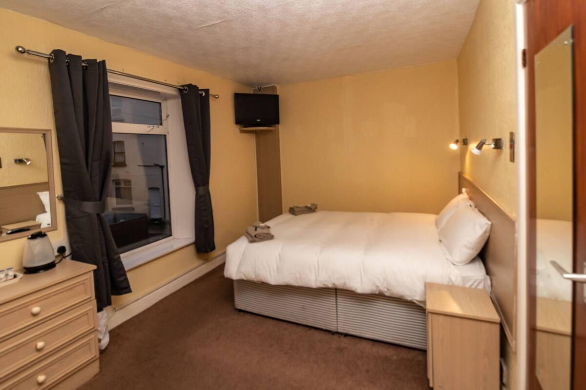 Un dormitorio con cama, cómoda, ventana con cortinas, televisor de pantalla plana montado en la pared y mesita de noche.