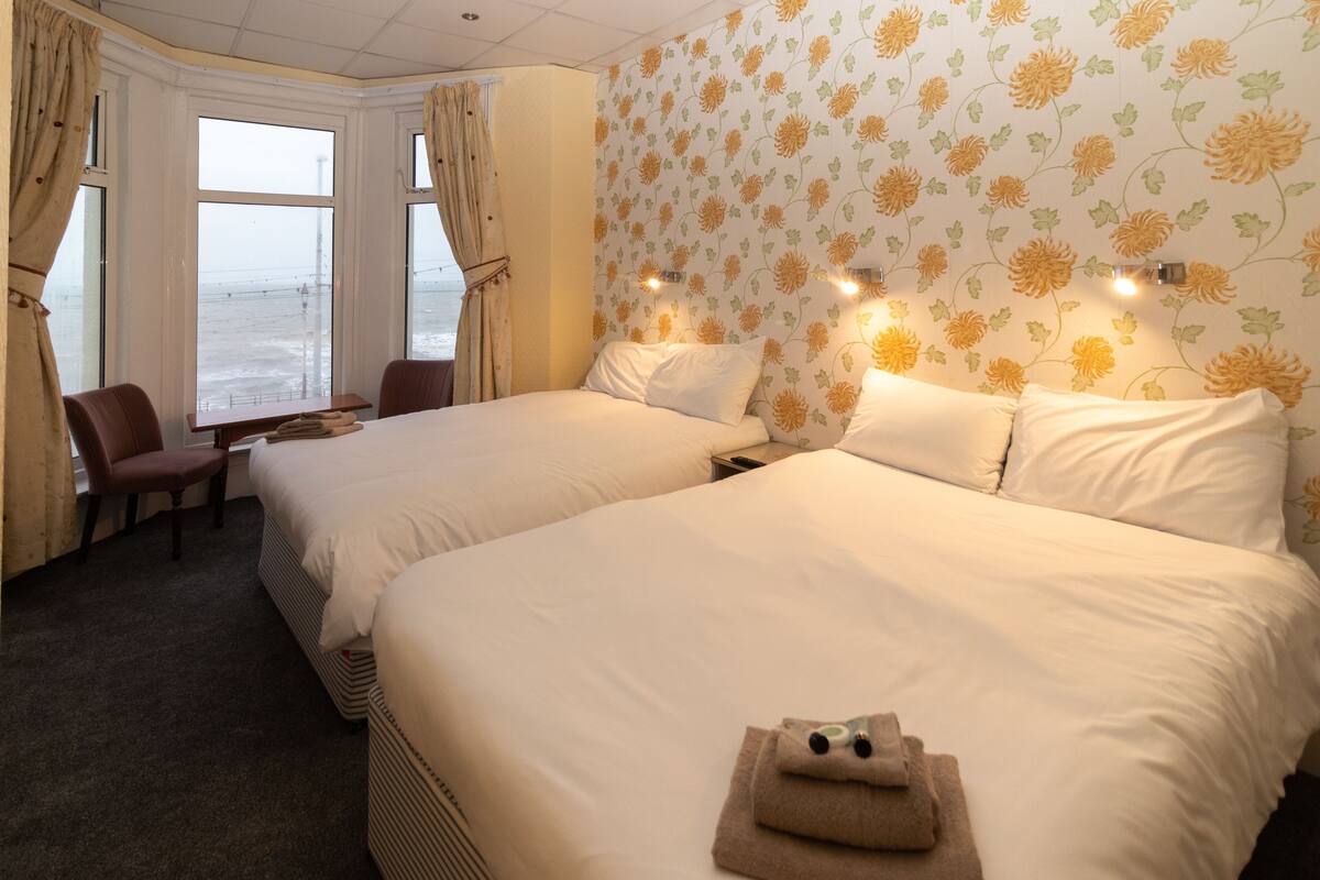 Una habitación de hotel con dos camas, un escritorio y una silla. Hay ventanas con cortinas, un papel tapiz floral y luces fijadas a la pared.