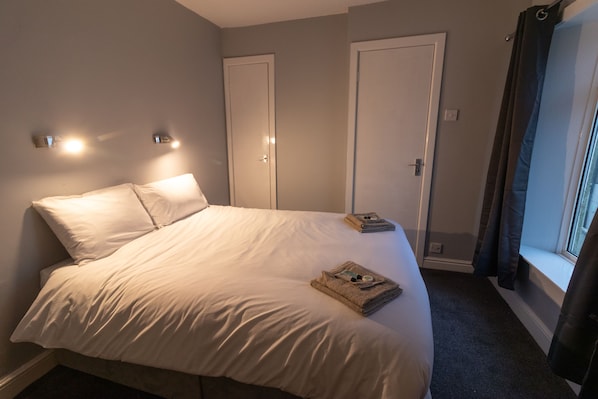 A neatly made bed with white linens, a folded towel, and a book on the bed. There are two doors and a window with curtains on the right side.