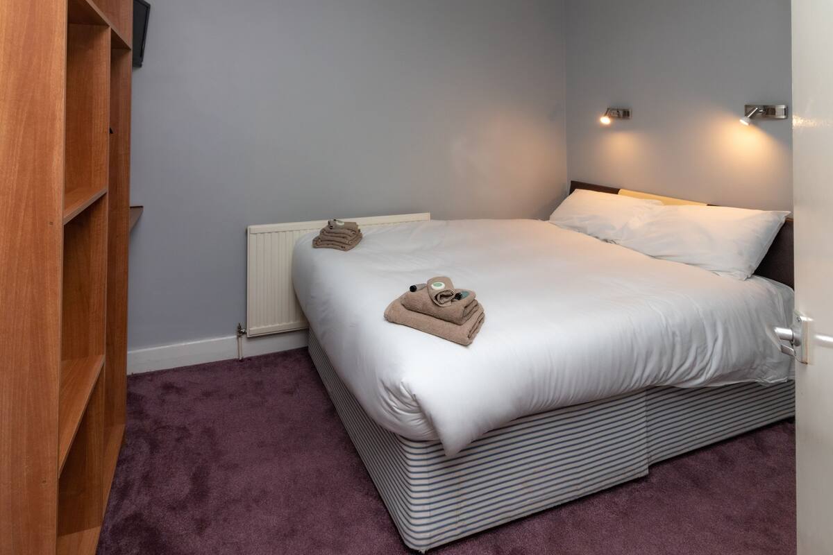 Una cama bien hecha con ropa de cama blanca y almohadas a rayas, un armario de madera y una alfombra morada.