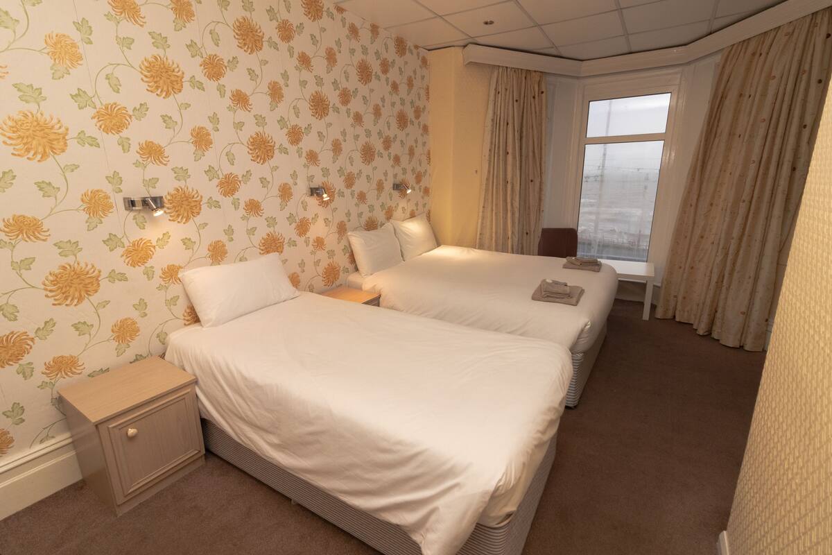 Habitación de hotel con dos camas, papel tapiz floral, una mesita de noche y una ventana con cortinas.