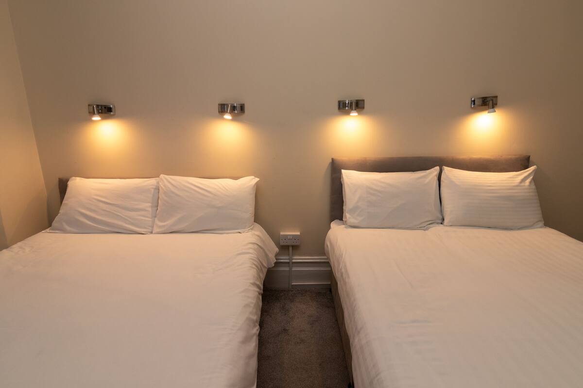 Habitación de hotel con dos camas, cada una con ropa blanca y cabeceros, y lámparas fijadas a la pared.