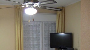 A small room with a ceiling fan, a television on a cabinet, a dining table with chairs, and a sliding glass door leading to a balcony.