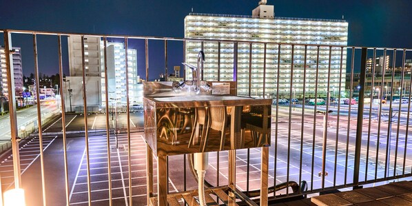A view from a balcony with a metal railing, overlooking a parking lot and a brightly lit building in the background.
