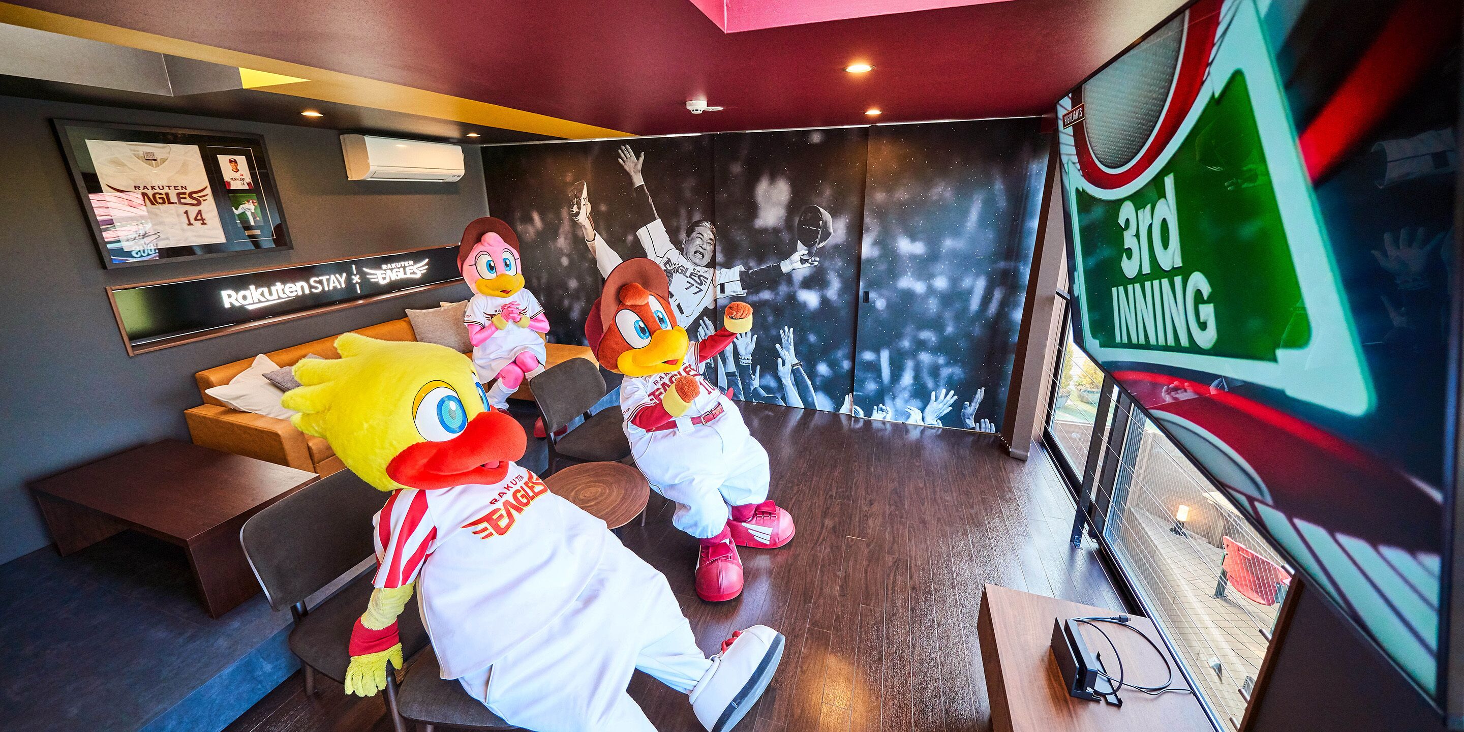 Ein Zimmer mit einem großen Bildschirm, der '3. INNING' anzeigt, und zwei Maskottchen. Ein Schild mit 'Rakuten Stay' und ein gerahmtes Trikot sind an der Wand zu sehen.