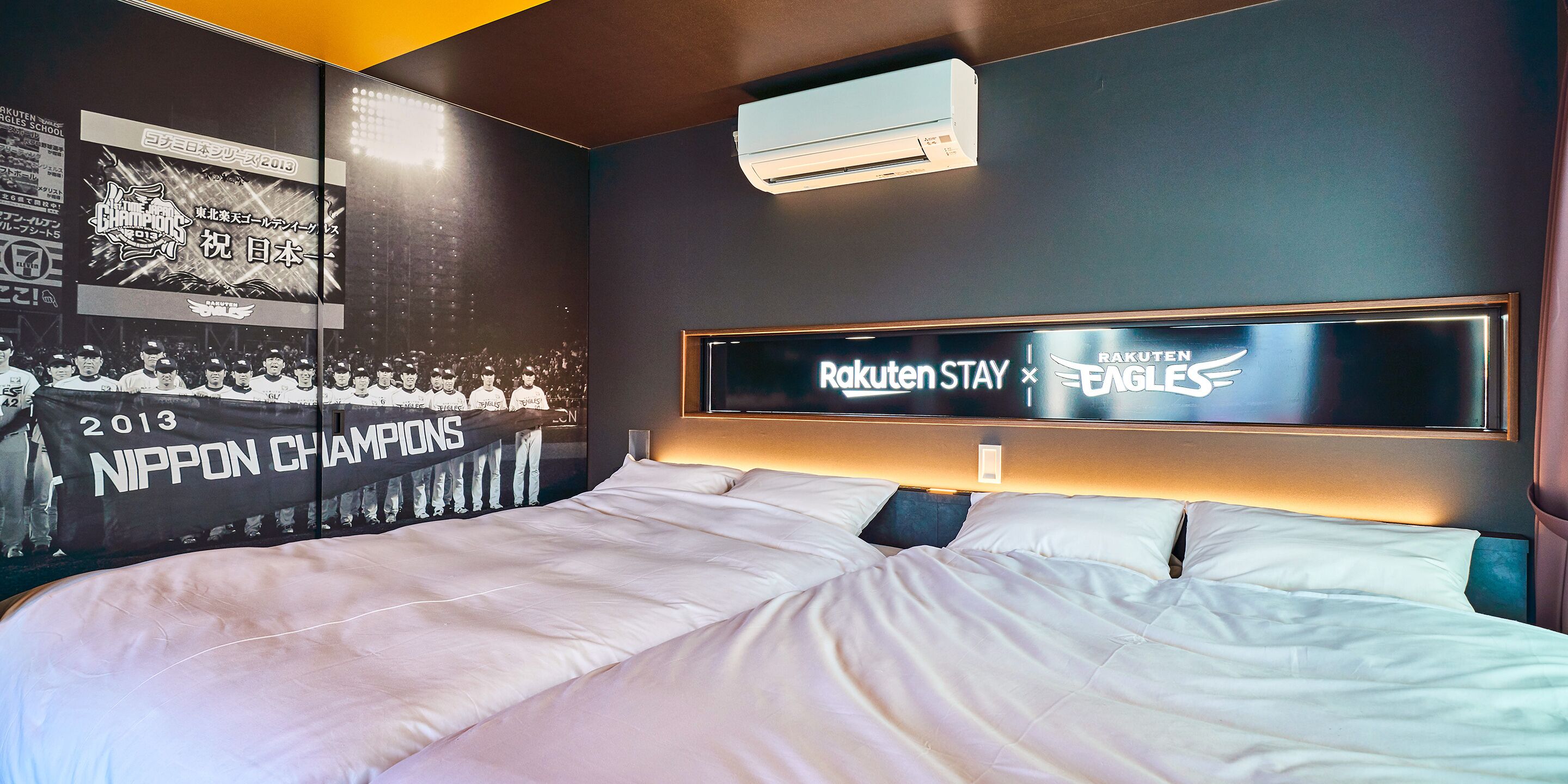 Une chambre d’hôtel avec un grand lit, un climatiseur fixé au mur et une affiche de l’équipe de baseball Nippon Champions, vainqueur de l’édition 2013.