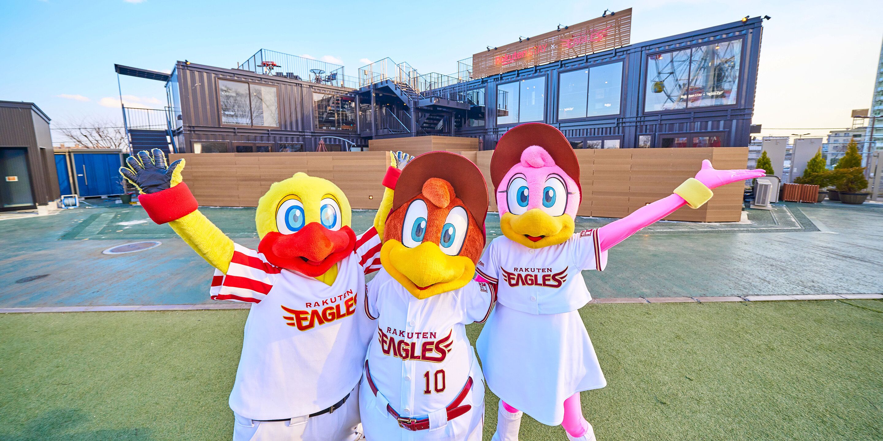 Tres mascotas con uniformes deportivos de pie en un campo de deportes, con edificios al fondo.