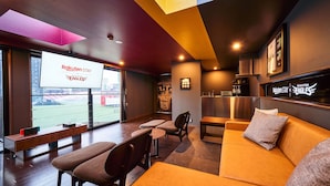Un área de descanso moderna con asientos, una pantalla grande mostrando 'Rakuten Stay Eagles' y vista a un campo deportivo por la ventana.