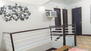 Una habitación con pared blanca, decoración floral en blanco y negro, unidad de aire acondicionado blanca, un banco de madera, una mesa con mantel floral y una escalera con barandilla de madera.