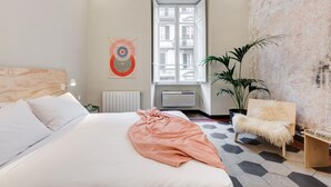 Ein modernes Schlafzimmer mit einem großen Bett, einer Topfpflanze, einem Sessel und einem Fenster mit Blick auf Gebäude.