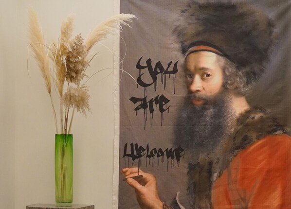 A person standing next to a wall with graffiti text and a green vase with dried plants.