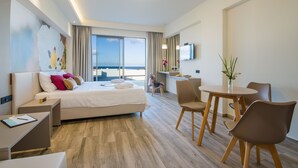 A modern hotel room with a large bed, bedside tables, a desk, a dining area with chairs, a small table with a vase, and a TV on the wall.