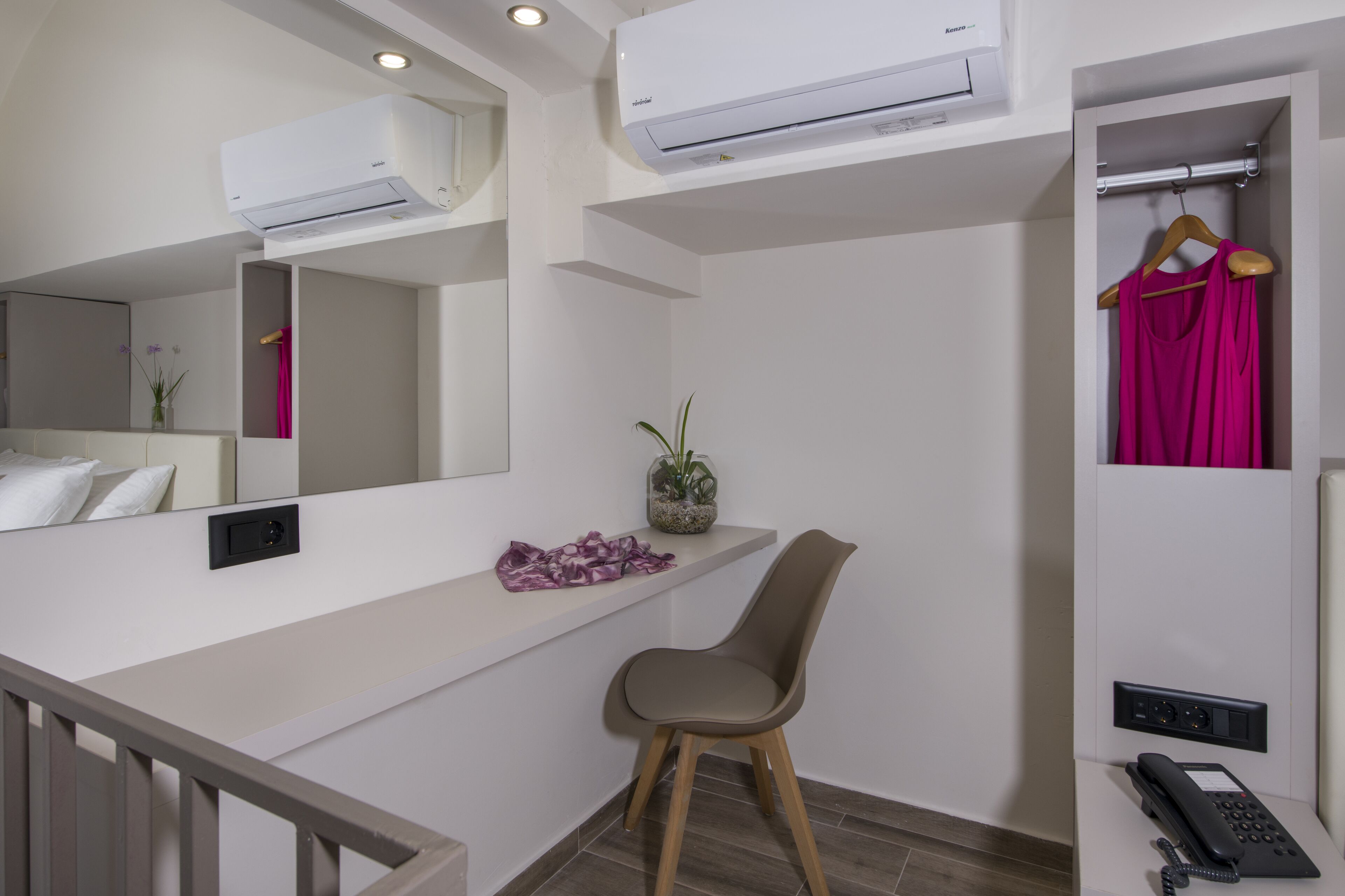 Une chambre d’hôtel moderne avec une coiffeuse blanche, un miroir, une petite plante, une chaise en bois, un peignoir et un téléphone sur une table de chevet.