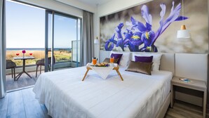 A hotel room with a large bed, a dining table with breakfast, a view of the beach, and a large floral painting on the wall.