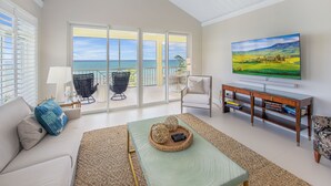 A spacious living room with a large TV, a sofa, a coffee table, and a view of the ocean through large sliding doors. - Ocean Front Luxury Condo Panoramic Views of the Caribbean Sea  (George Town)