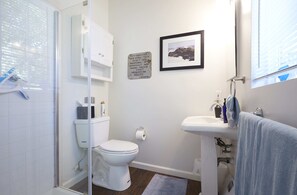 A bathroom with a toilet, sink, and shower. There is a framed picture on the wall and a sign with text.
