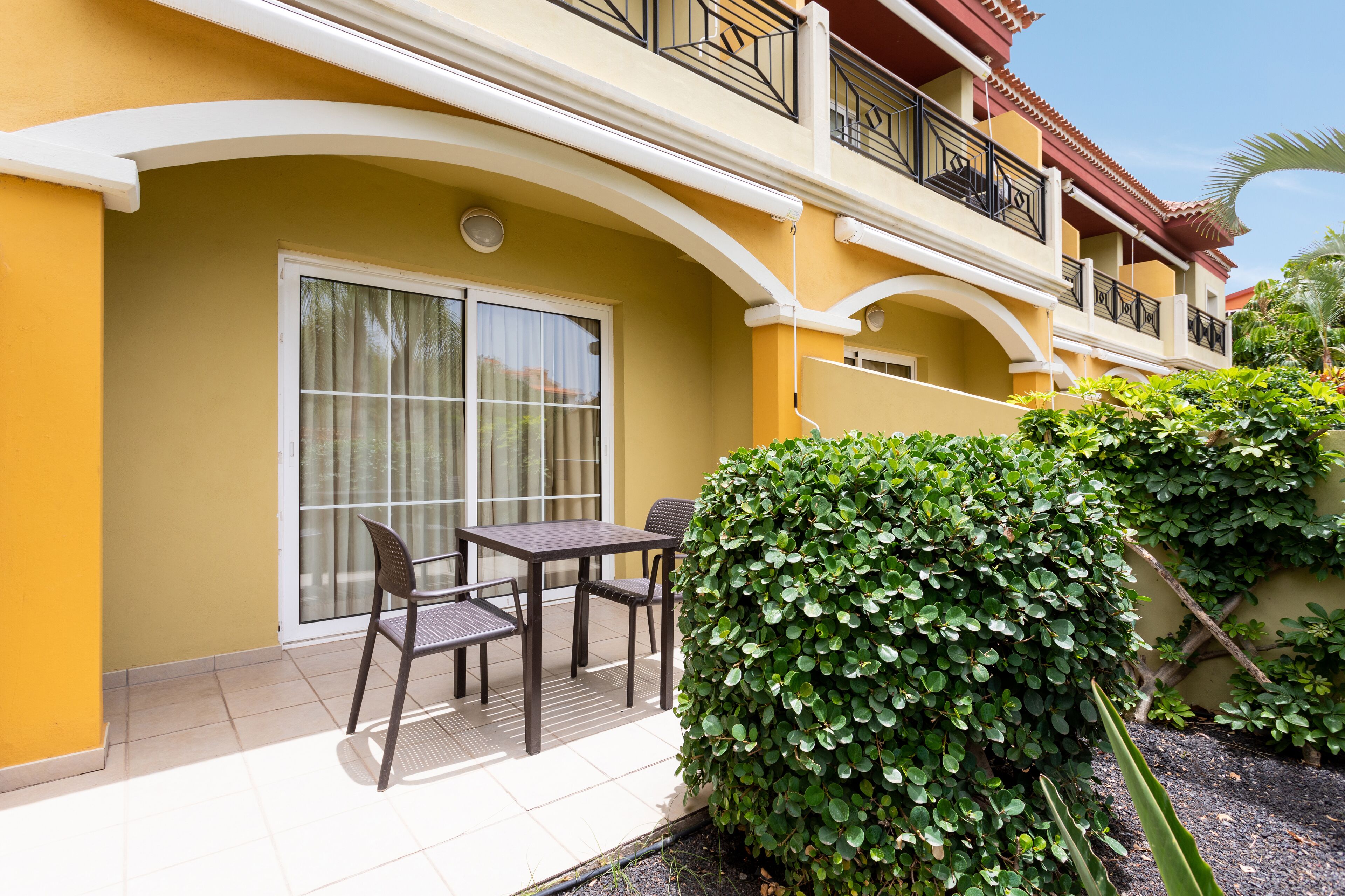 A yellow building with a balcony, a table with chairs, and a well-maintained hedge.