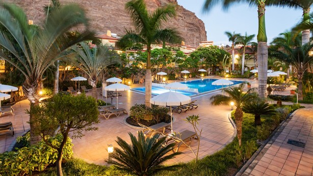 A resort pool area with lounge chairs and umbrellas, surrounded by palm trees and a large rock formation.