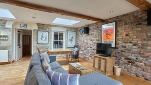 Un vaste espace de vie comprenant un grand canapé, une table basse en bois et un téléviseur fixé sur un mur en briques. Des tableaux encadrés ornent le mur, et une lucarne se trouve au-dessus de l’espace détente.