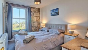 A bedroom with a large bed, a stone wall, a view of the outdoors, and a painting on the wall.