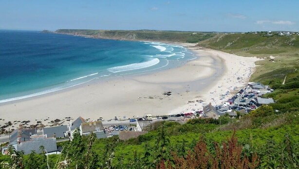 A coastal beach with a sandy shore, a clear blue sea, and a few buildings along the coastline.