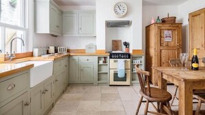 A kitchen with light green cabinets, a wooden table with chairs, a clock, and a wooden cabinet with a sign. - Gin O'Clock, Mevagissey, Level Walk to Harbour! (Mevagissey)
