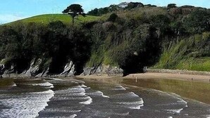A coastal landscape with a sandy beach, a rocky shoreline, and a grassy hill with trees. - Gin O'Clock, Mevagissey, Level Walk to Harbour! (Mevagissey)