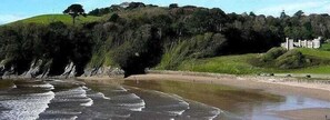 Un paisaje costero con una playa arenosa, un litoral rocoso y una colina cubierta de pasto y árboles.