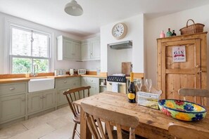 A kitchen with light green cabinets, a wooden table with a bottle of champagne, a colorful bowl, and a clock on the wall. - Gin O'Clock, Mevagissey, Level Walk to Harbour! (Mevagissey)
