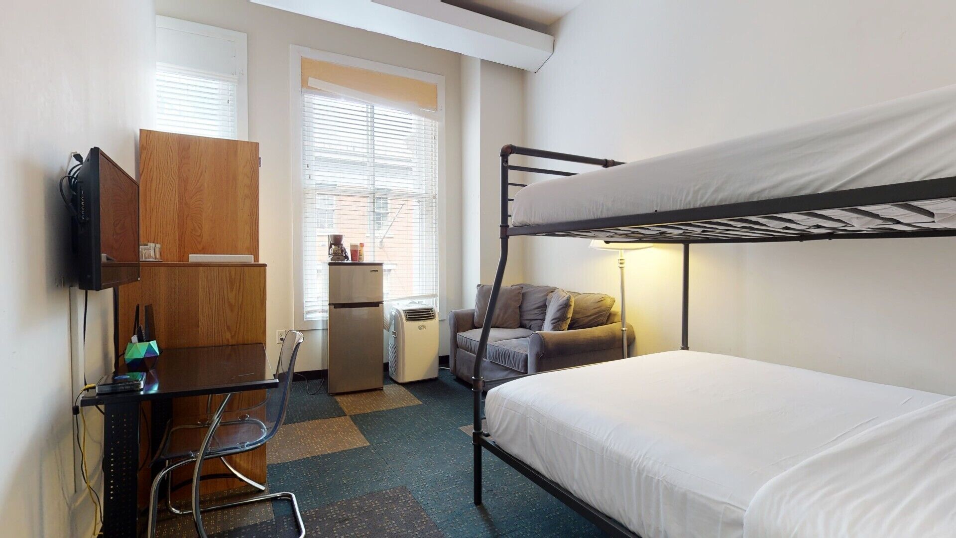 Een compacte hotelkamer met een stapelbed, een bureau met stoel, een televisie, een koelkast en een raam met jaloezieën.