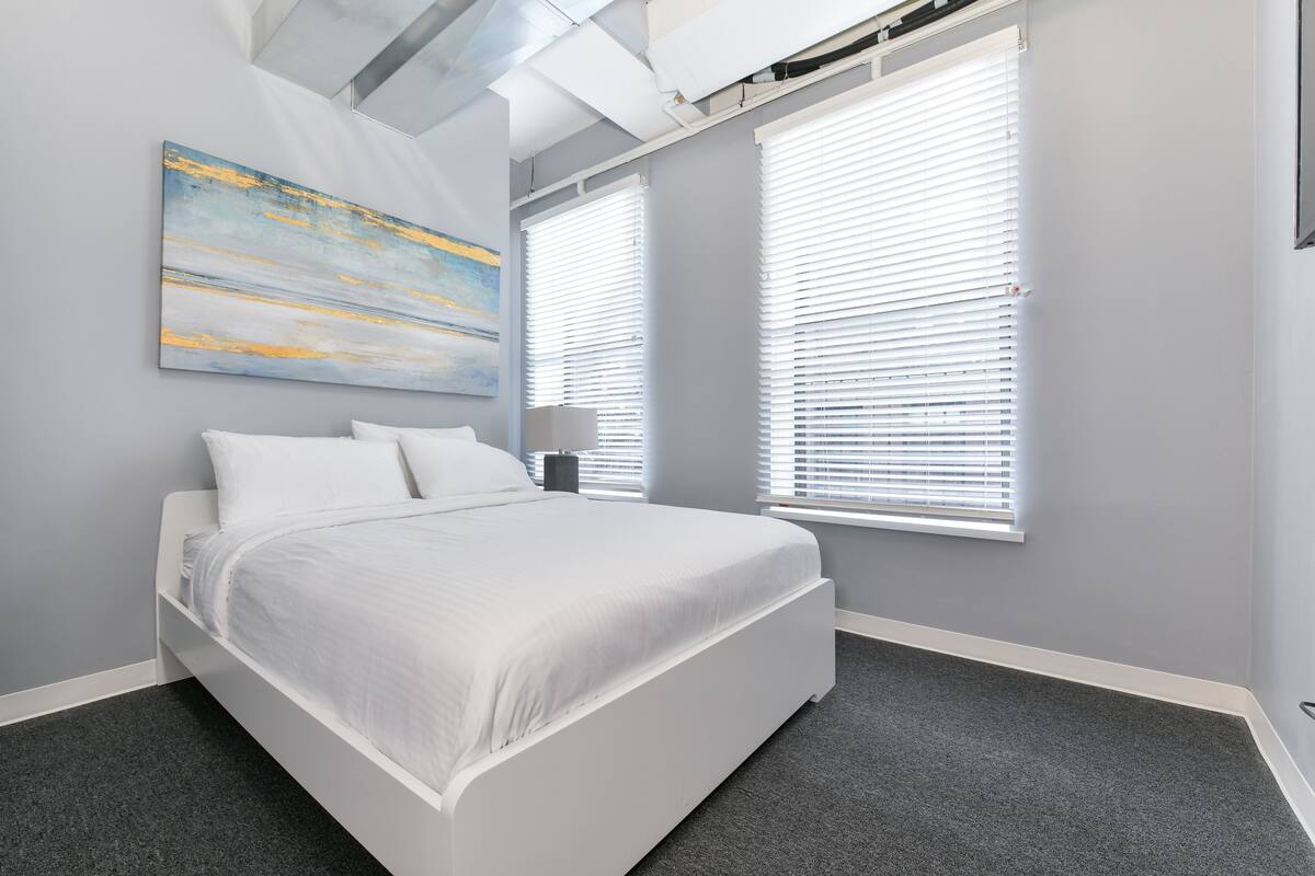 Un dormitorio con cama, dos ventanas con persianas, un cuadro en la pared y techo con conductos a la vista.