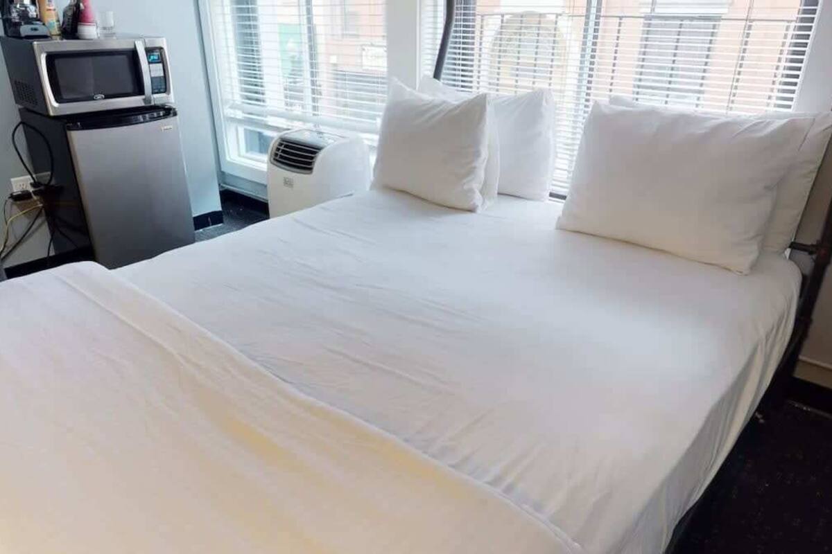 Una habitación de hotel con una cama grande, ropa de cama blanca, un escritorio pequeño con una computadora y una ventana con persianas.