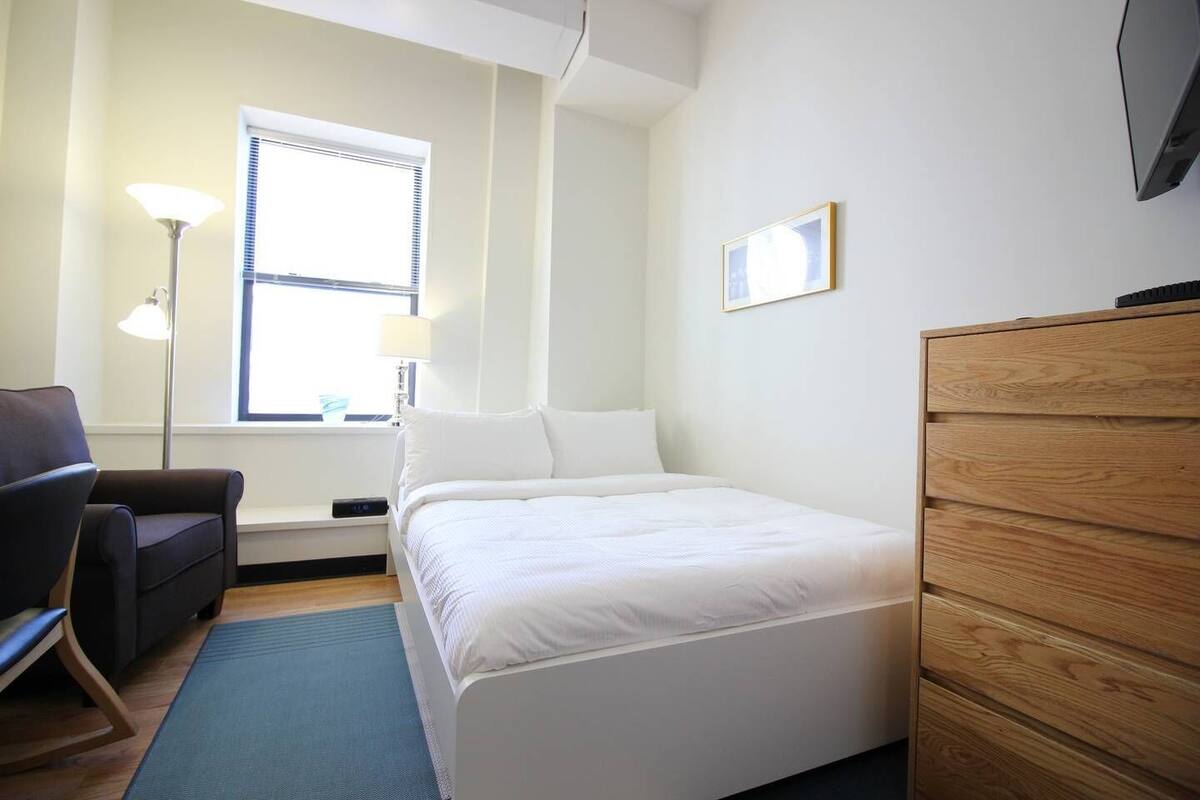 Un dormitorio con cama, cómoda, silla, lámpara y ventana.