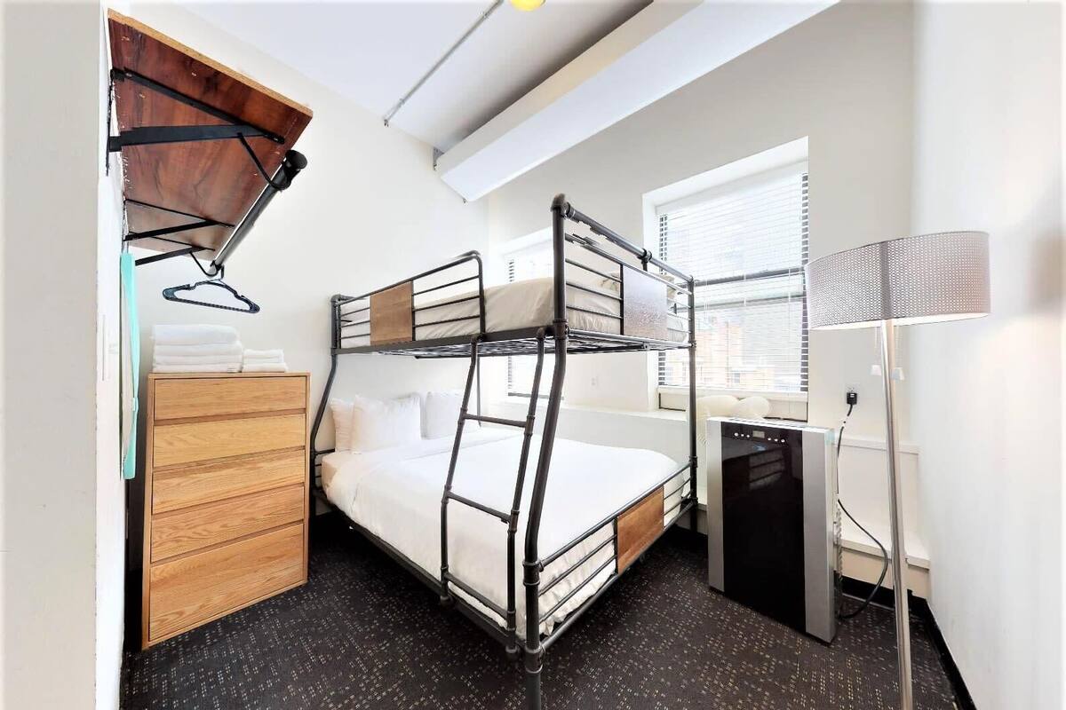 Habitación de hotel compacta con literas, cómoda de madera, escritorio con computadora y ventana con persianas.