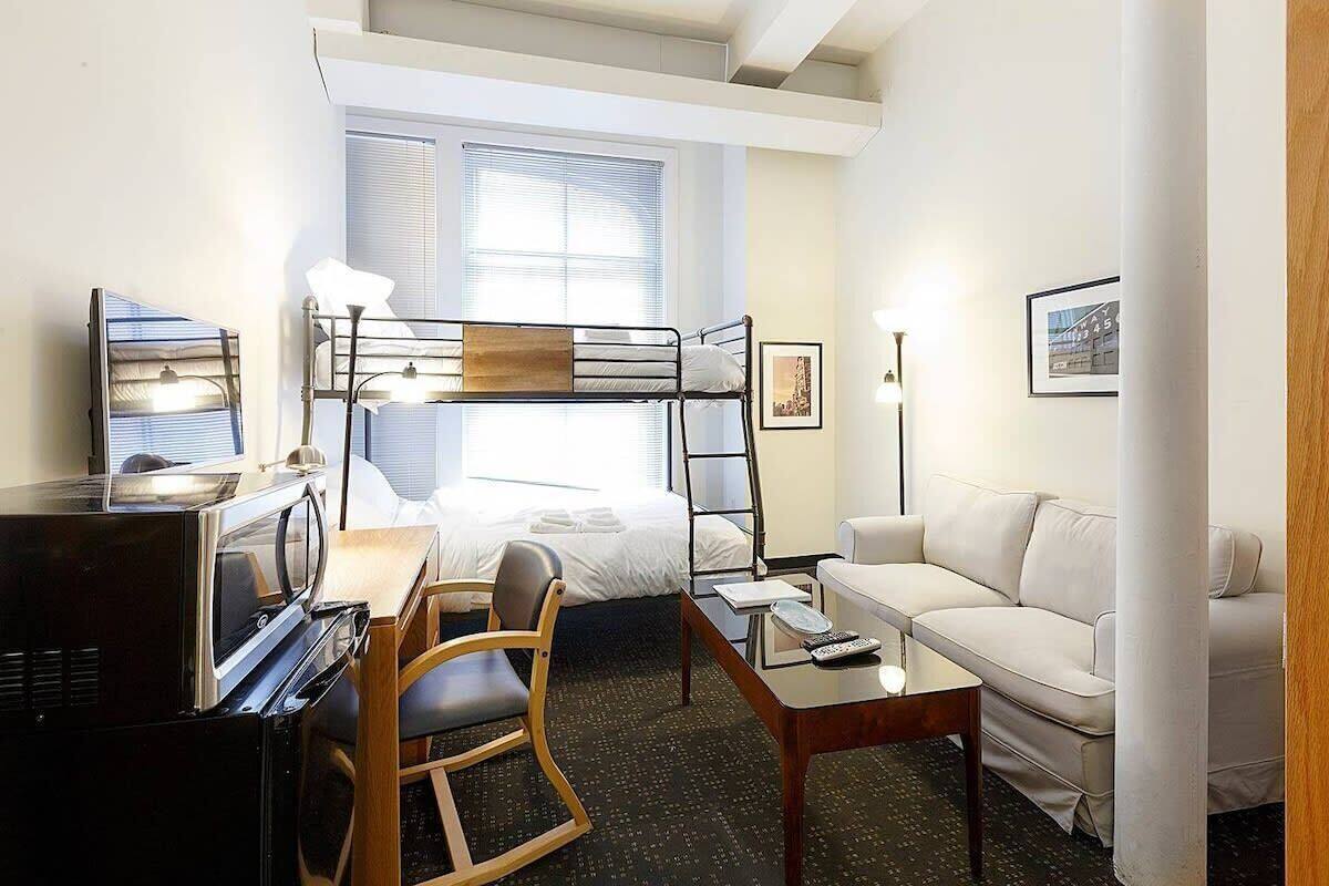 Una habitación de hotel compacta con una cama individual superior, un escritorio con ordenador, una silla, un sofá y una mesita de centro.