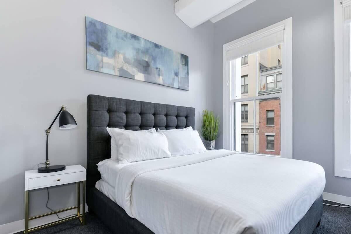 Un dormitorio moderno con una cama grande, una mesita de noche con lámpara, una ventana con vistas a edificios y un cuadro en la pared.