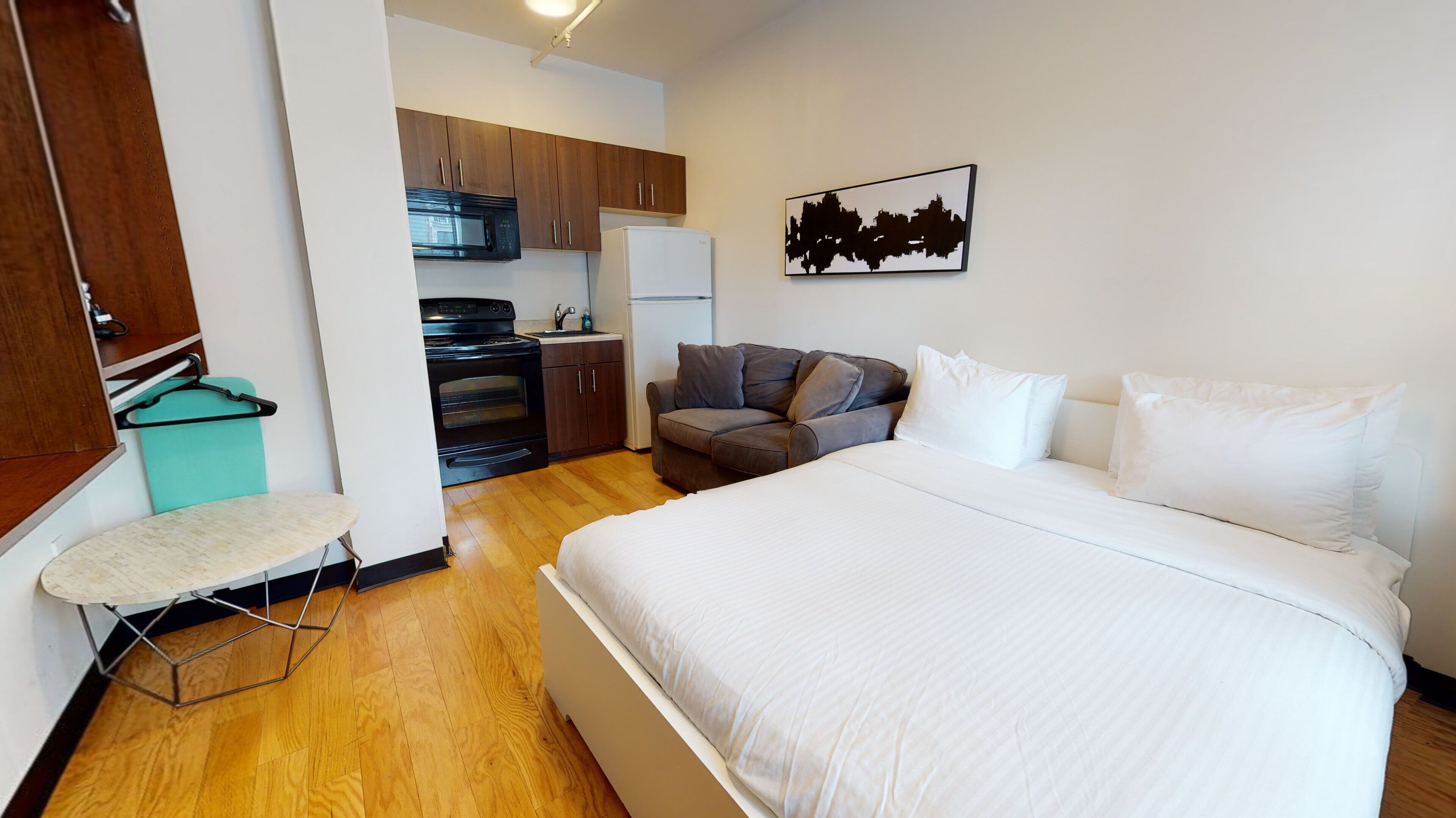 A bedroom with a large bed, a grey sofa, a kitchen area with a microwave and oven, and a wall-mounted artwork.